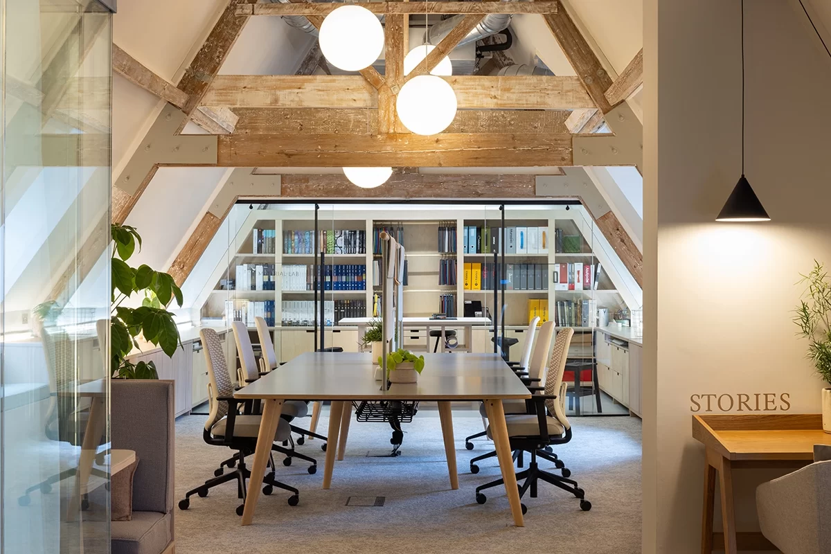 OP Birmingham office upstairs area with big table surrounded by beige task chairs and wooden slabs with 4 round pendant lights. At the back there's the design area with designers samples and books.